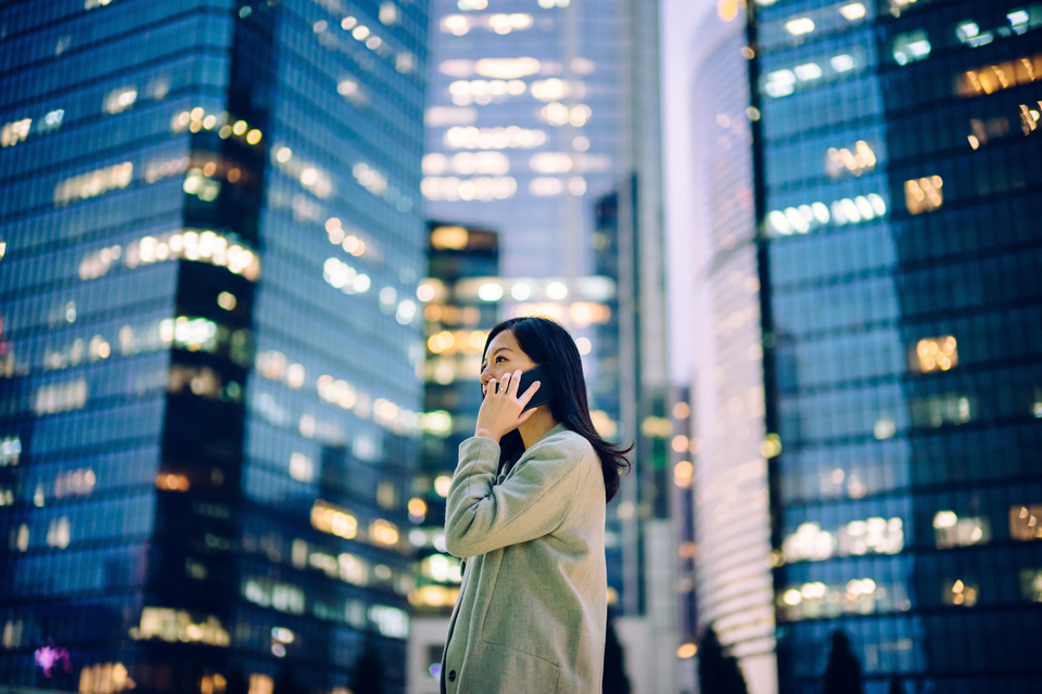 Young woman with mobile phone standing in front of business buildings with shining lights | © © 2019 Yiu Yu Hoi