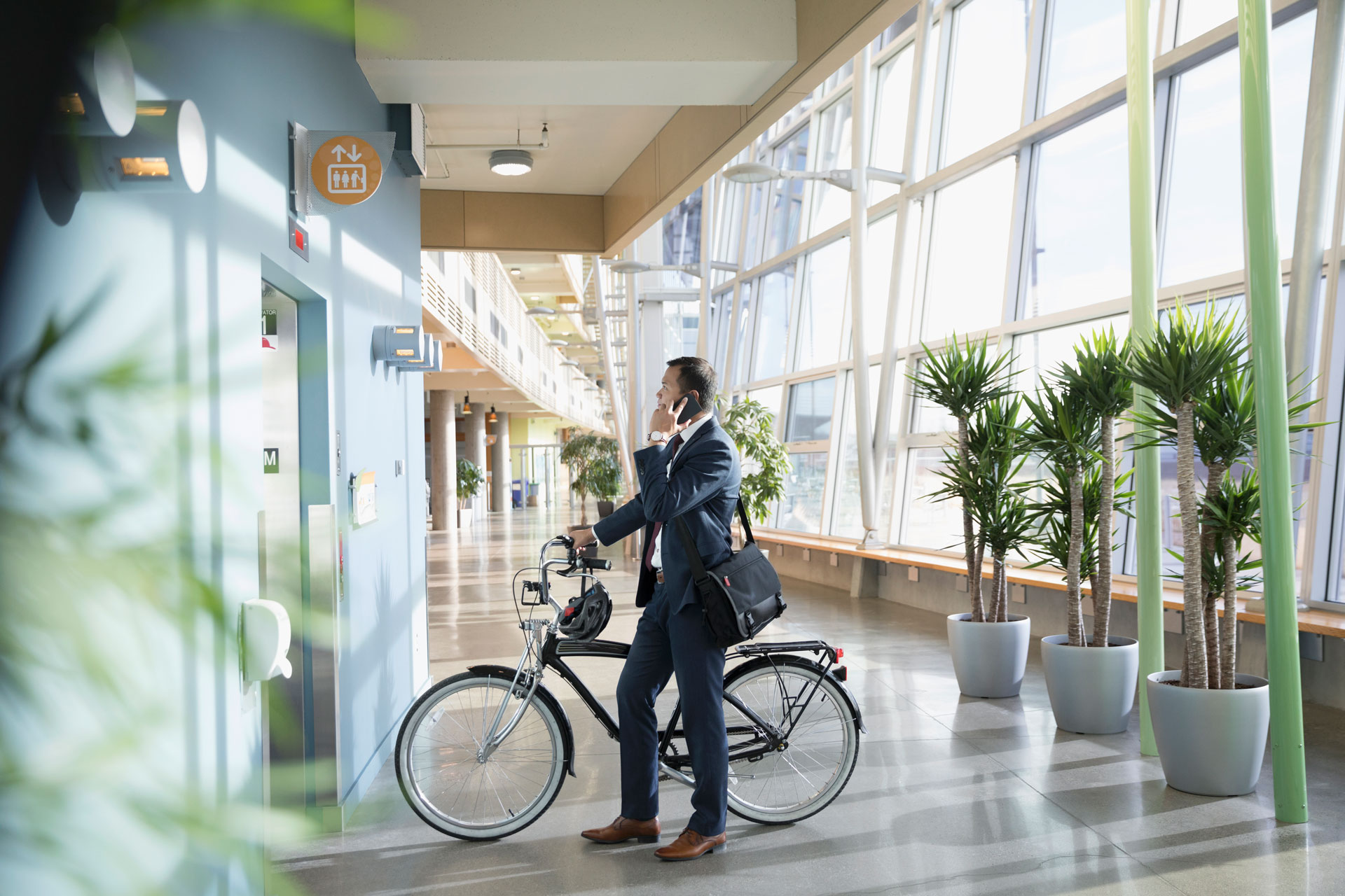 Businessman commuter with bicycle talking on smart phone, waiting at elevator in office lobby