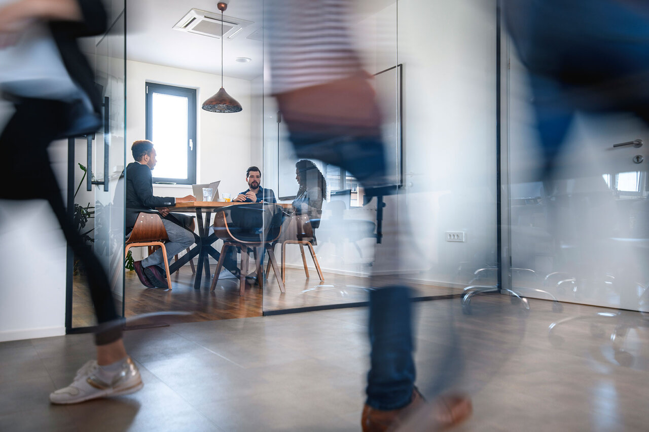 Blurred motion of colleagues walking briskly down office hallway as colleagues sit in conference room discussing ideas. | © AzmanL @GettyImages